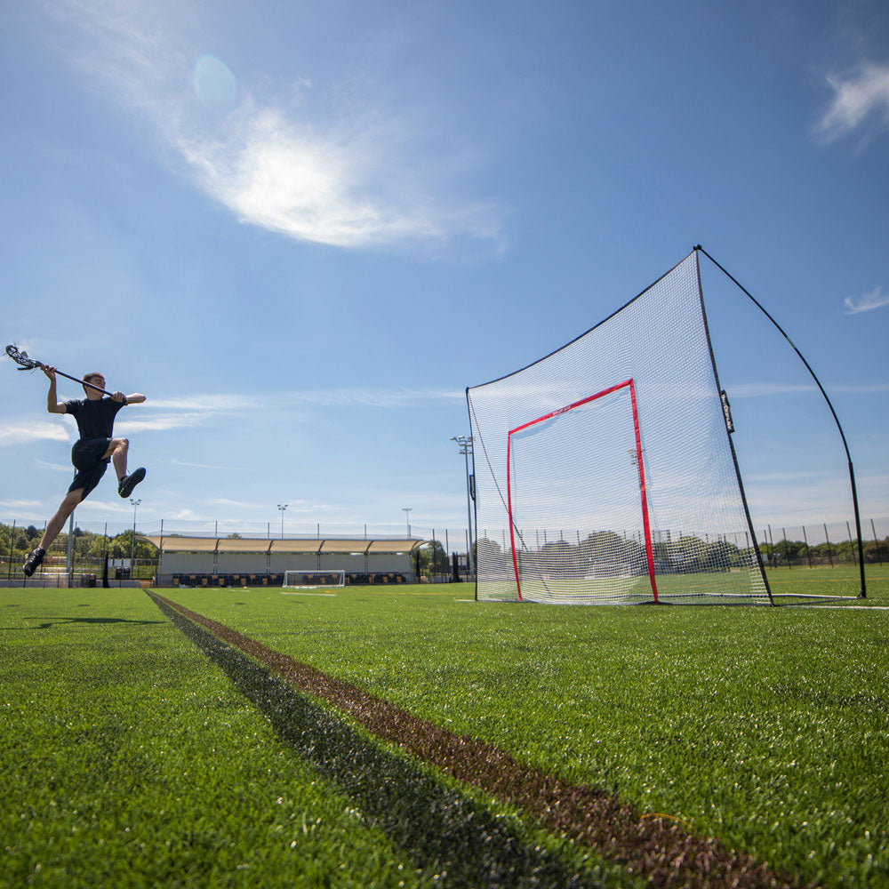 Lacrosse-Spieler springt hoch, um auf ein MEGA NET Element mit orangefarbenem Zielrahmen auf einem Kunstrasenplatz zu schießen.
