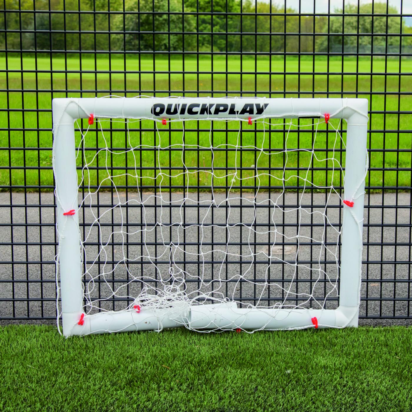 Q-FOLD Mini-Fußballtor 90 x 75 cm flach zusammengeklappt auf Kunstrasen vor einem schwarzen Gitterzaun.