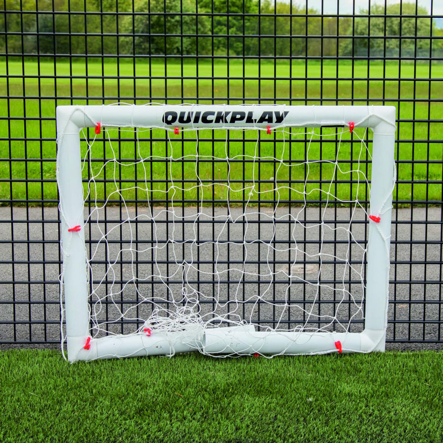 Q-FOLD Mini-Fußballtor 90 x 75 cm flach zusammengeklappt auf Kunstrasen vor einem schwarzen Gitterzaun.