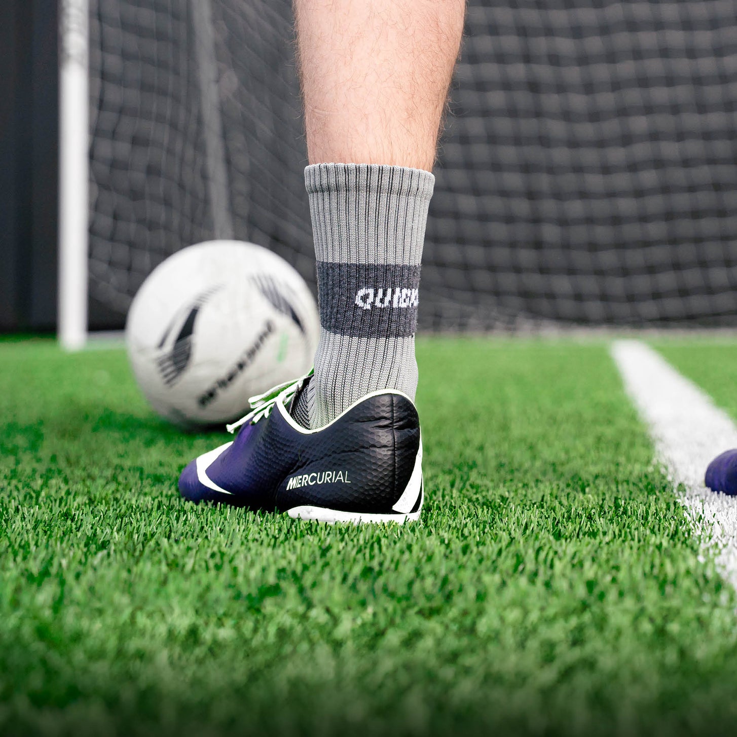 Spieler trägt eine graue QUICKPLAY Grip-Socke mit Fußballschuh auf Kunstrasen, dahinter Ball und Tornetz.