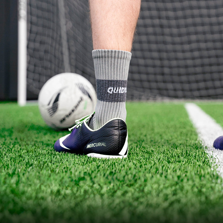 Spieler trägt eine graue QUICKPLAY Grip-Socke mit Fußballschuh auf Kunstrasen, dahinter Ball und Tornetz.