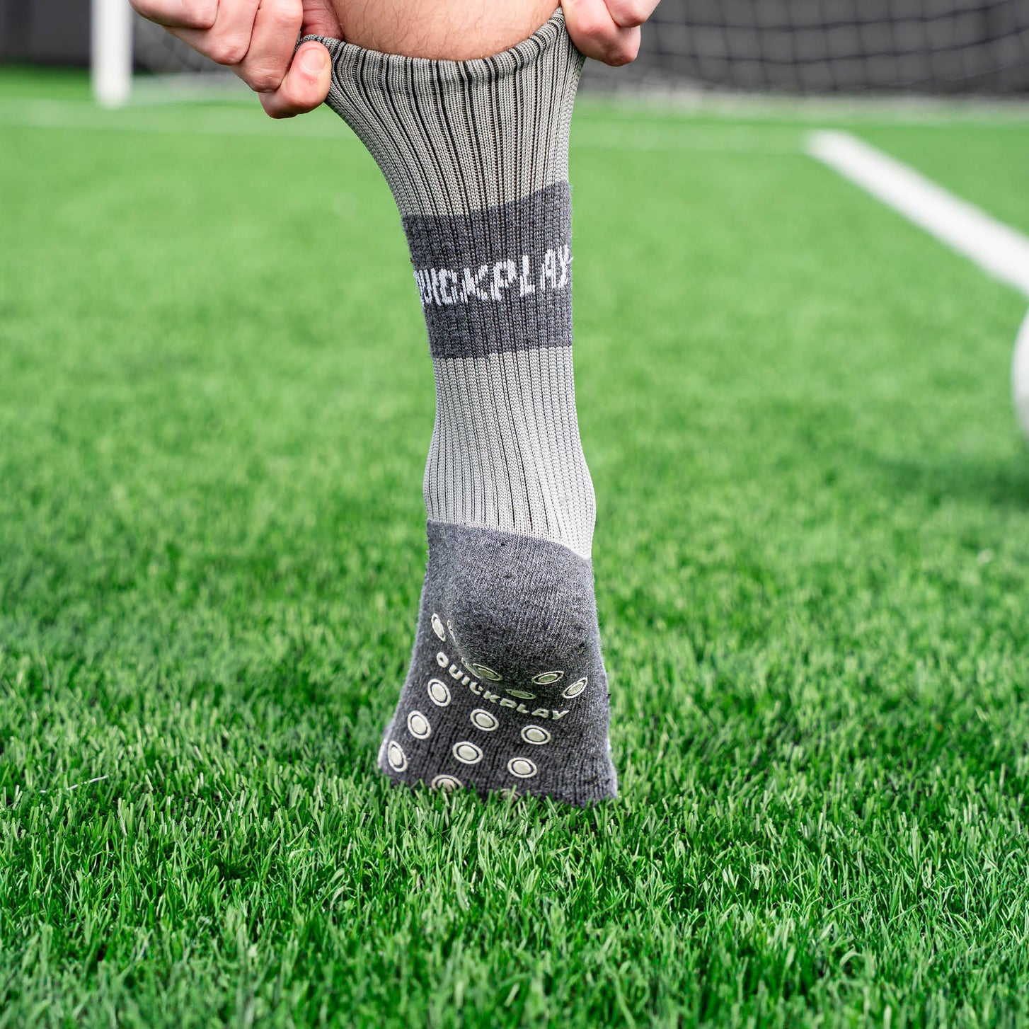 Spieler zieht auf Kunstrasen eine QUICKPLAY Grip-Socke hoch, die Sohle mit Grip-Noppen zeigt zur Kamera.