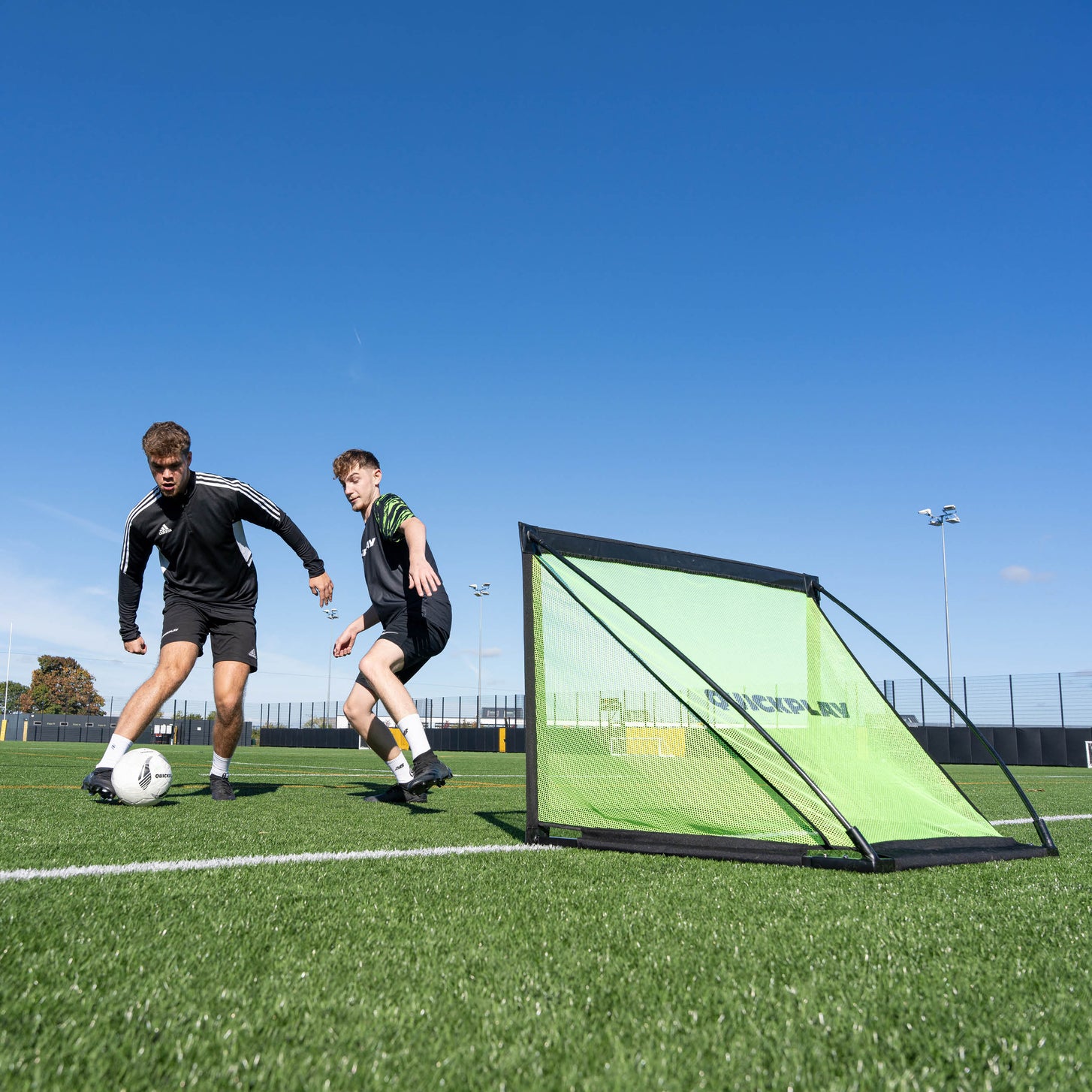 Spieler dribbeln an einem sonnigen Tag auf einem Kunstrasenplatz am KICKSTER PRO Tor 120 x 75 cm vorbei.