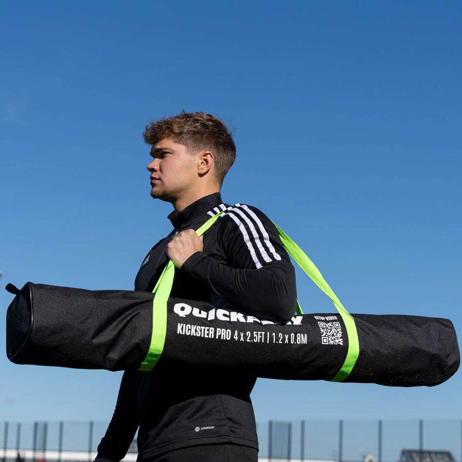 Spieler trägt eine schwarze QUICKPLAY KICKSTER PRO Tasche an den grünen Gurten über der Schulter, vor blauem Himmel.