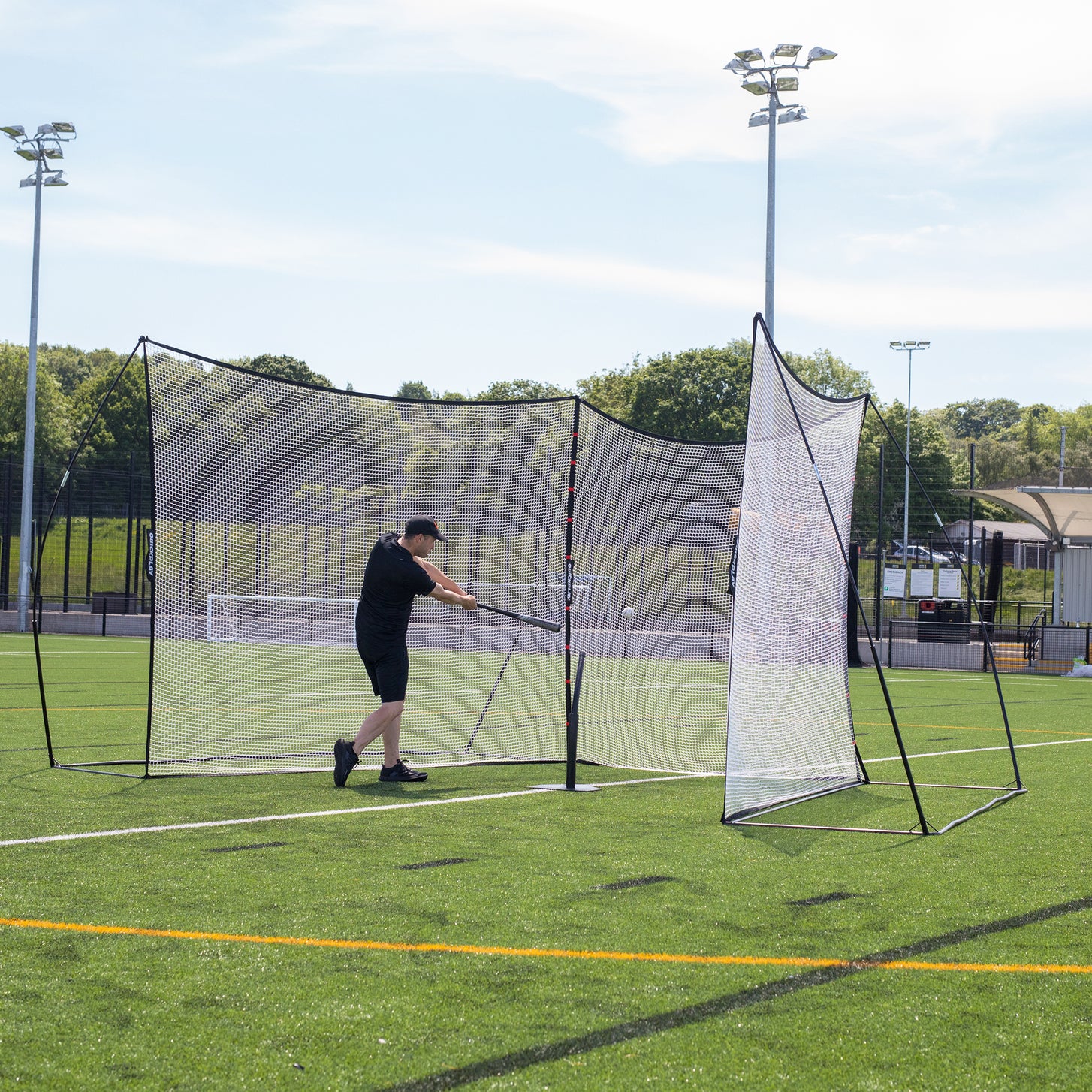 Mann schlägt mit einem Baseballschläger einen Ball in zwei MEGA NETs, verbunden durch das Erweiterungsnetz 240 x 270 cm, eine schwarze Tasche zu seinen Füßen.
