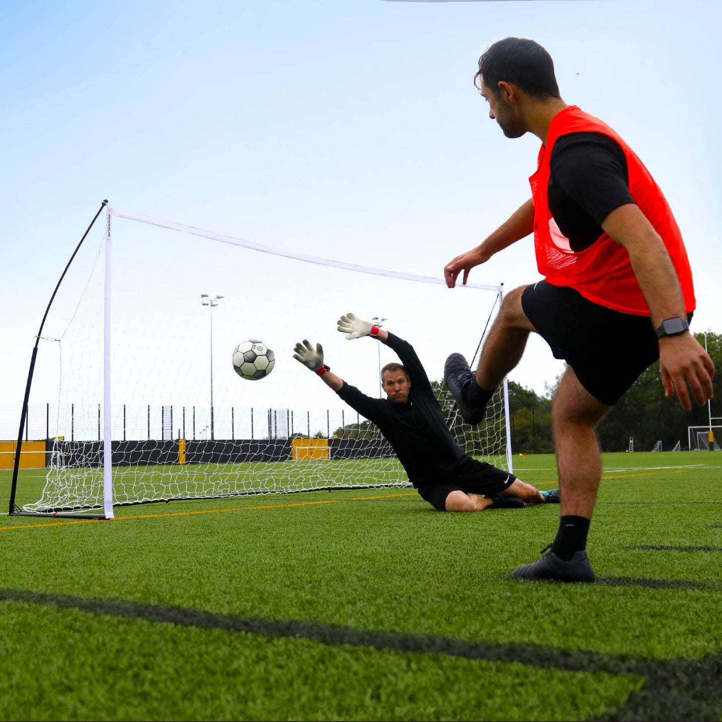 KICKSTER Elite Tor 370 x 180 cm auf Platzhöhe, ein Torwart streckt sich nach dem Schuss eines Spielers im orangefarbenen Leibchen.