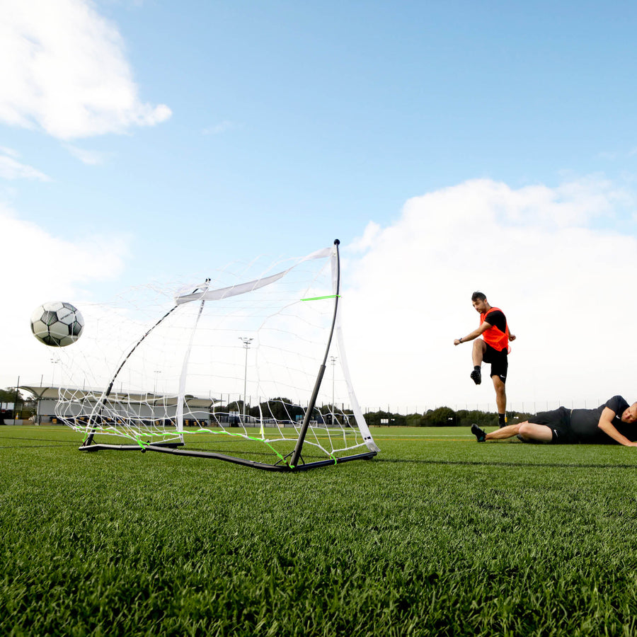 Aus niedriger Perspektive: Spieler schießt einen Ball am KICKSTER Elite Tor 150 x 100 cm vorbei, während ein anderer Spieler auf dem Platz liegt.
