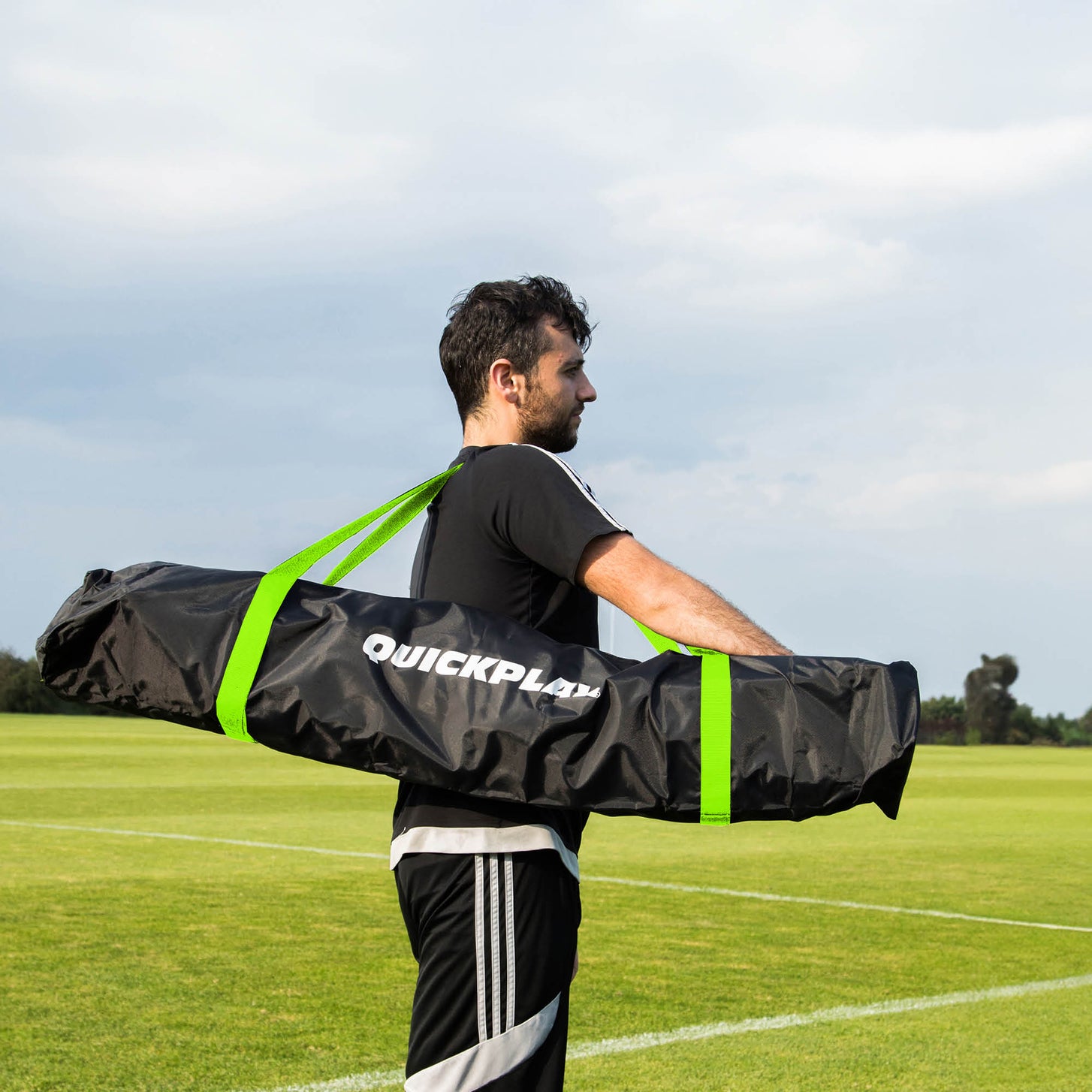 Mann trägt das MEGA NET 370 x 270 cm in seiner schwarzen QUICKPLAY Tasche mit grünen Gurten über ein Rasenfeld.