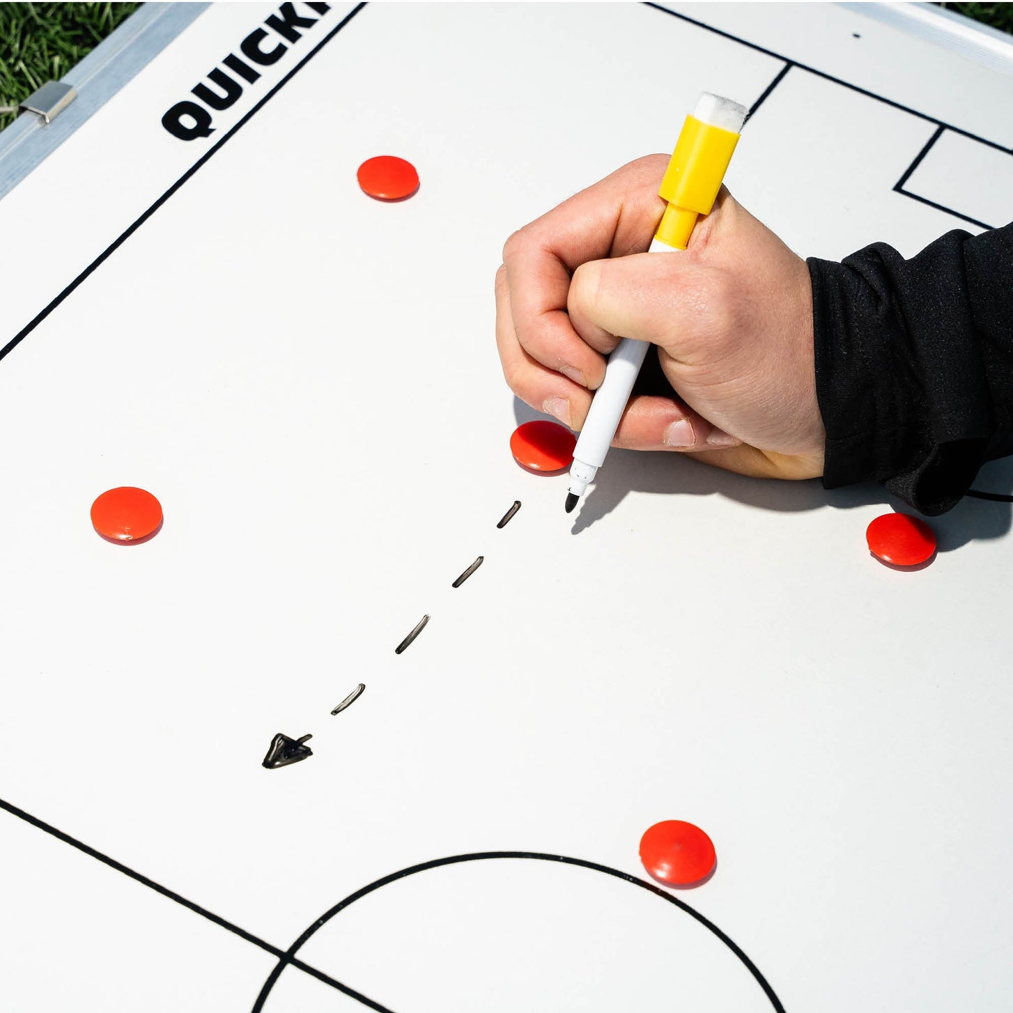 Magnetische Fußball-Taktiktafel 60 x 90 cm im Einsatz, eine Hand zeichnet mit gelbem Stift einen gepunkteten Pfeil zwischen roten Magneten.